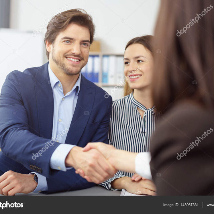 Smiling young couple shaking hands with a broker or agent as the man reaches across the table with a pleased friendly smile